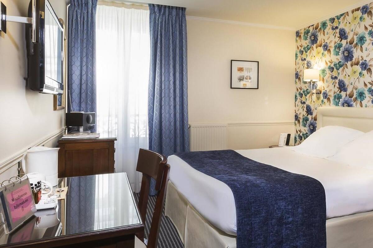 A hotel room with a bed, a desk, a chair, a TV, and a window with curtains.