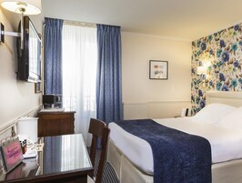Et hotelværelse med seng, skrivebord, stol, fjernsyn og et vindue med gardiner.