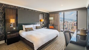 Habitación de hotel con una cama grande, dos mesitas de noche con lámparas, una silla, una pequeña mesa con un pastel, y un balcón con vistas a la ciudad.