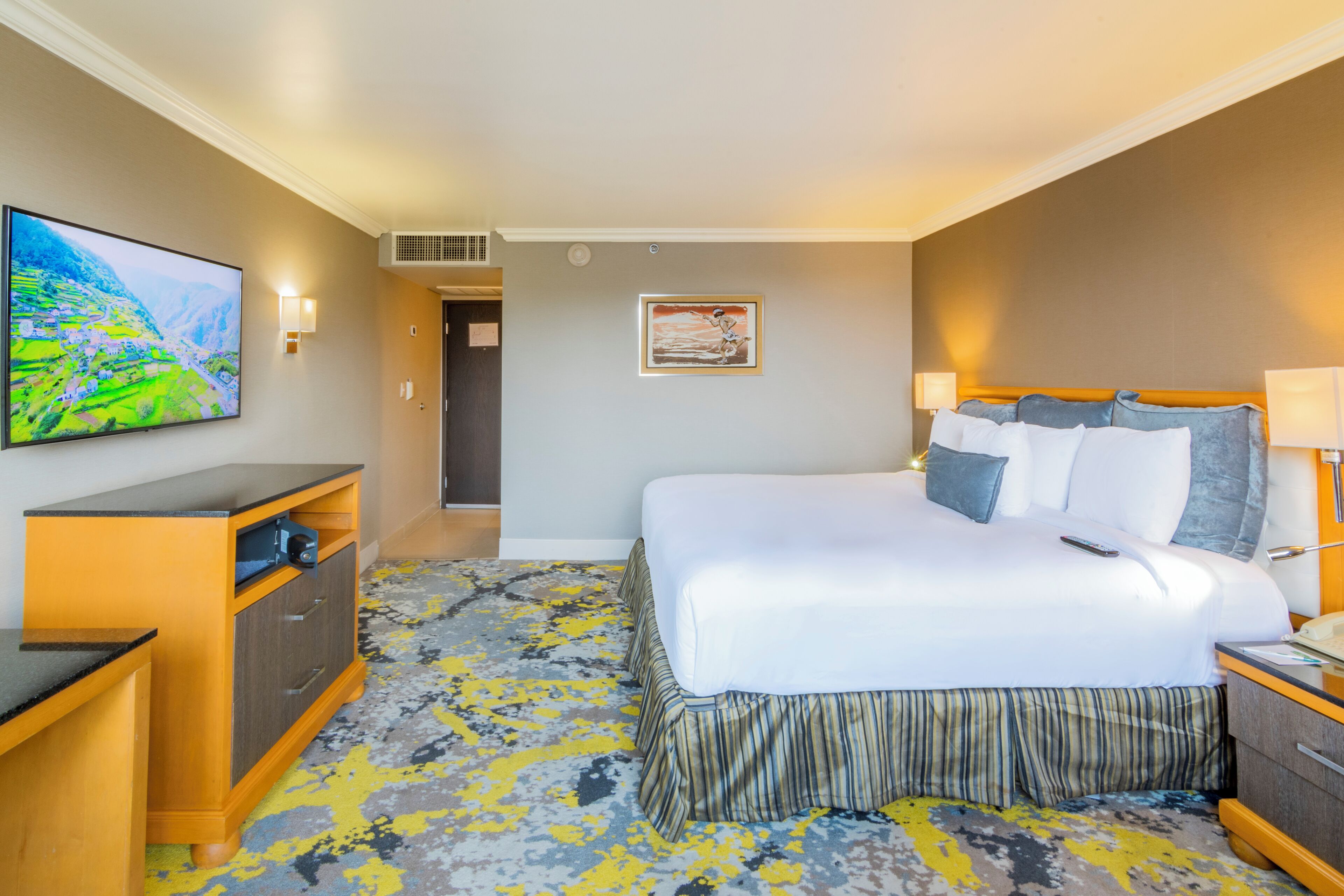A hotel room with a large bed, a TV mounted on the wall, a desk with drawers, and a carpeted floor.