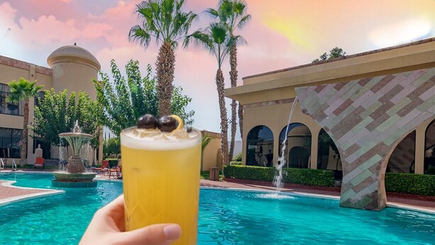 A hand holding a yellow drink with a garnish, a swimming pool, palm trees, and a building with arches in the background. - Hotel Lucerna Ciudad Juarez (Ciudad Juárez)