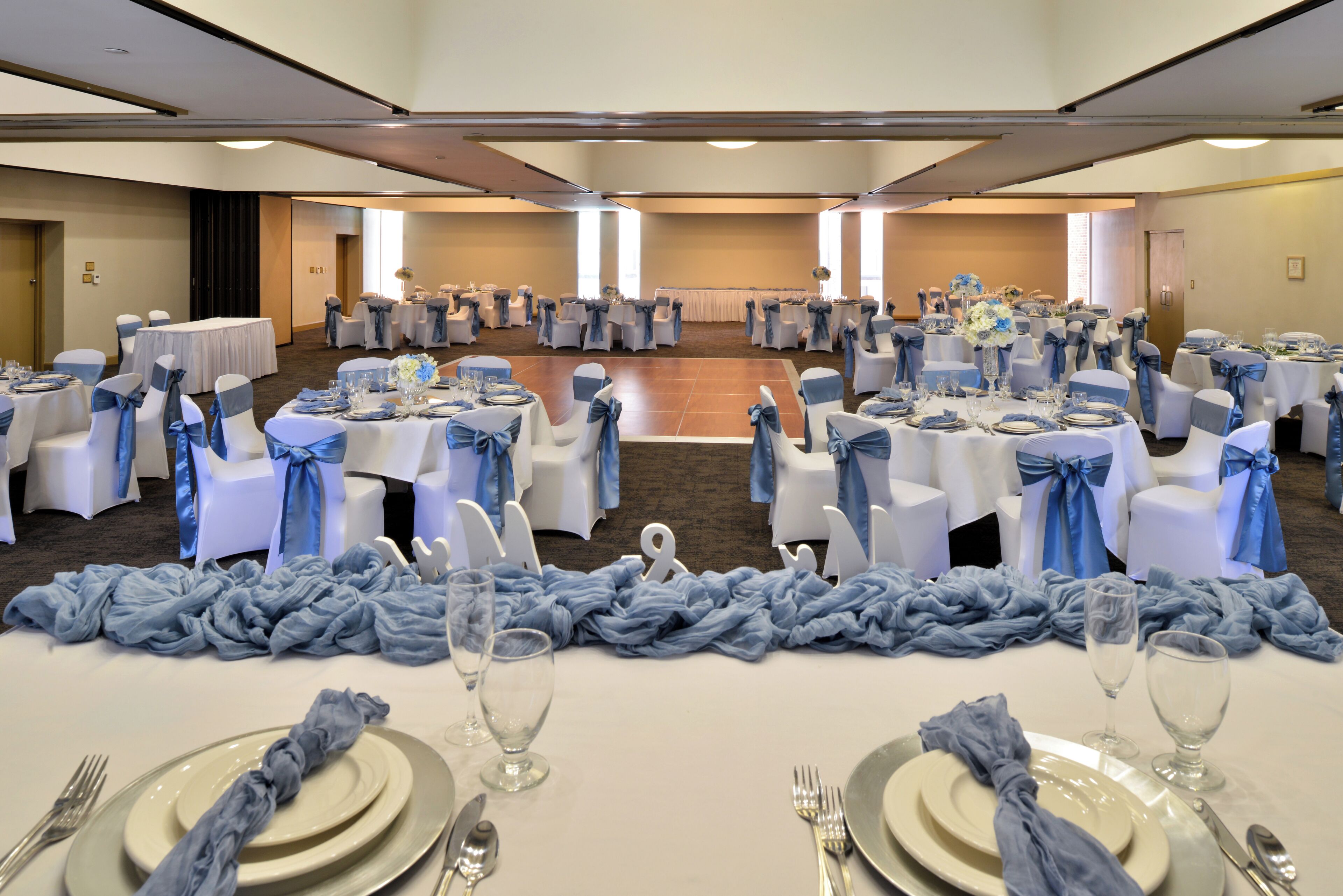 Un salón de banquetes con mesas redondas dispuestas para un evento formal, con sillas blancas y cintas azules, y suelo alfombrado.