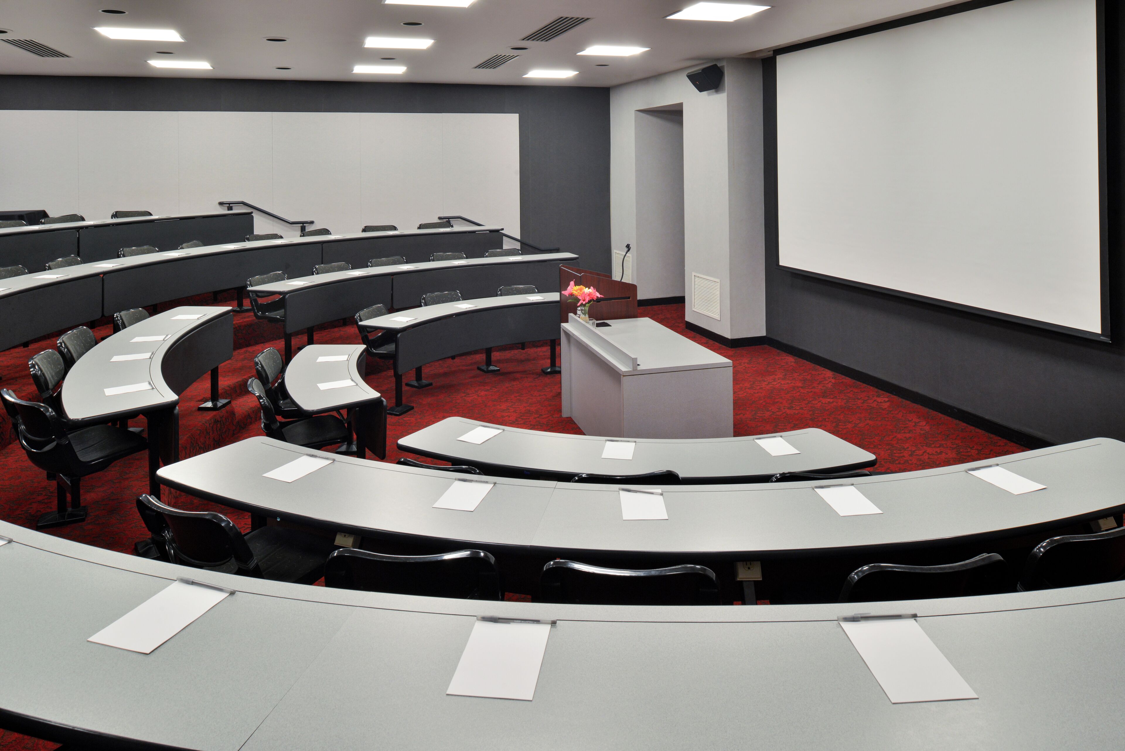 Sala de conferência com disposição em curva, várias mesas e uma tela de projeção grande.