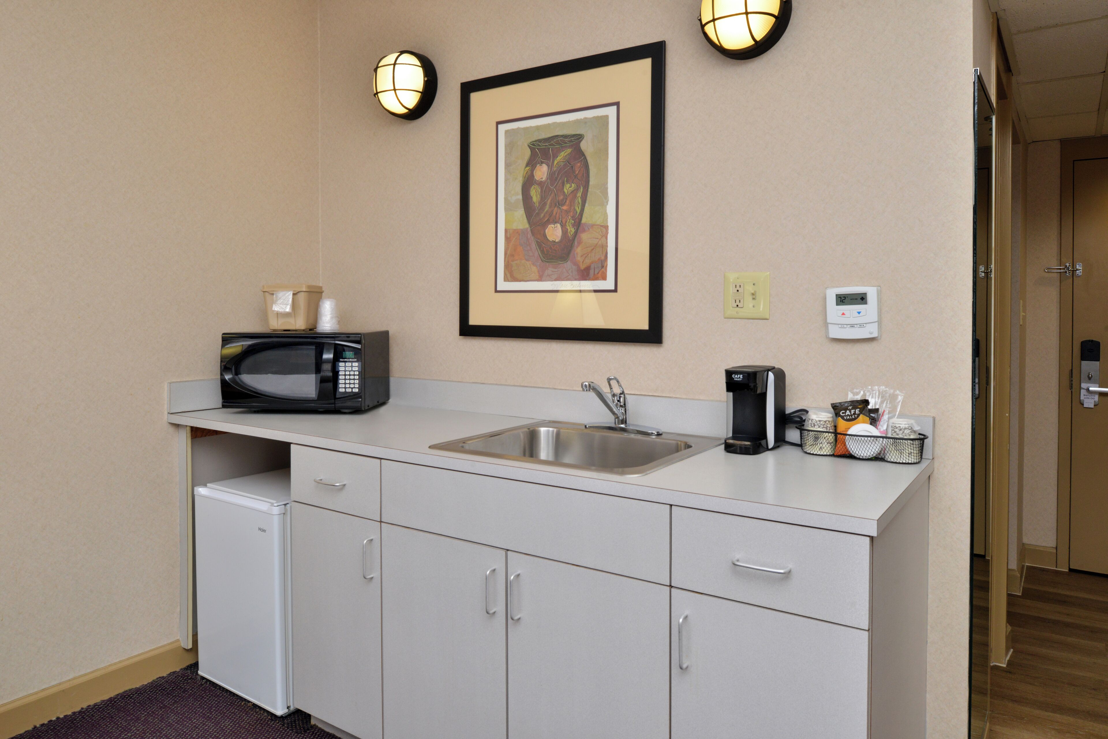 A kitchenette with a microwave, sink, and cabinets. There is a framed picture on the wall and a coffee maker on the counter.