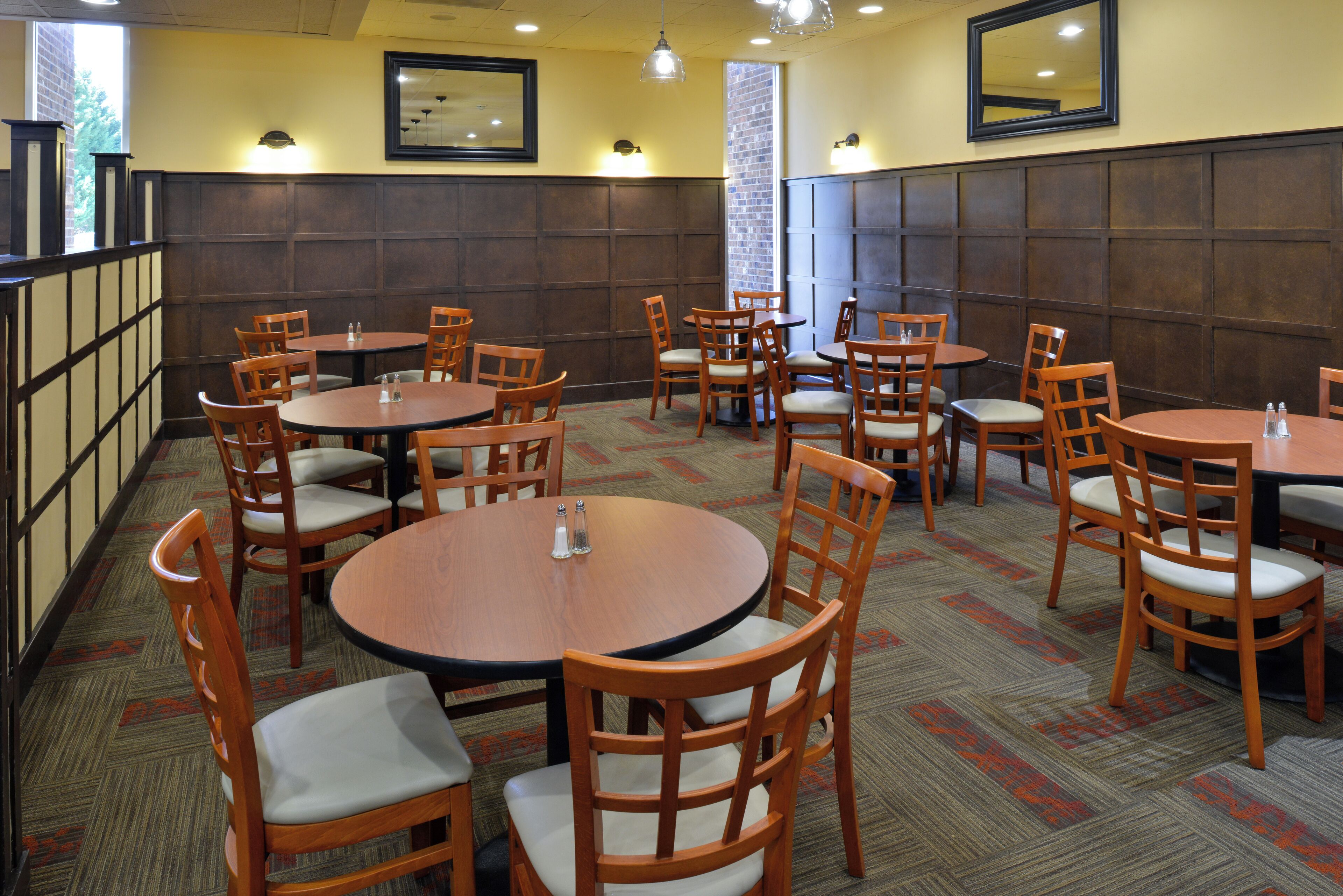A restaurant with multiple round tables and wooden chairs, arranged in a spacious layout with a high ceiling and wall-mounted lights.