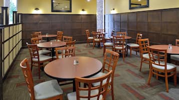A restaurant with multiple round tables and wooden chairs, arranged in a spacious layout with a high ceiling and wall-mounted lights.