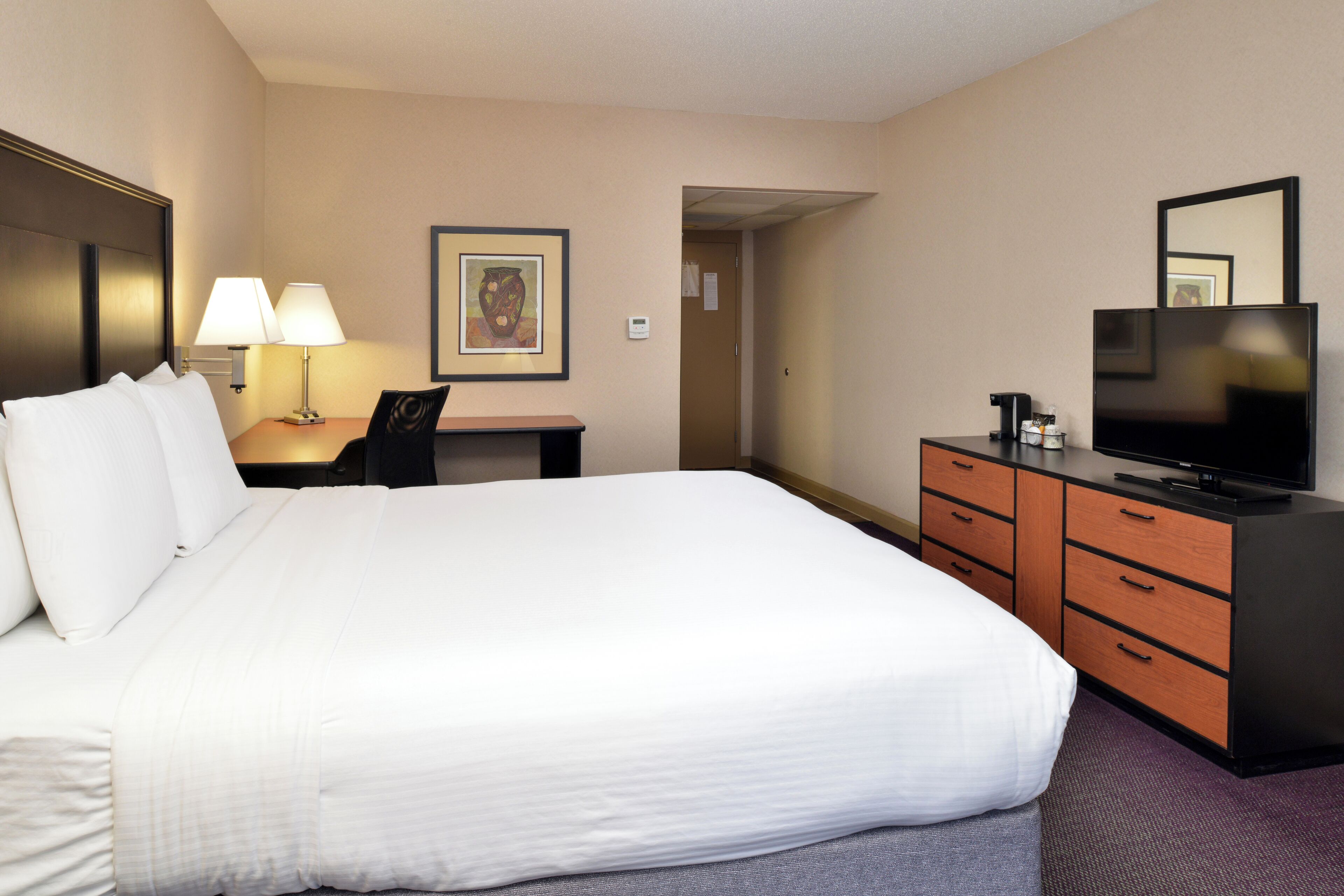 A hotel room with a large bed, a desk with a lamp, a television, and a dresser.
