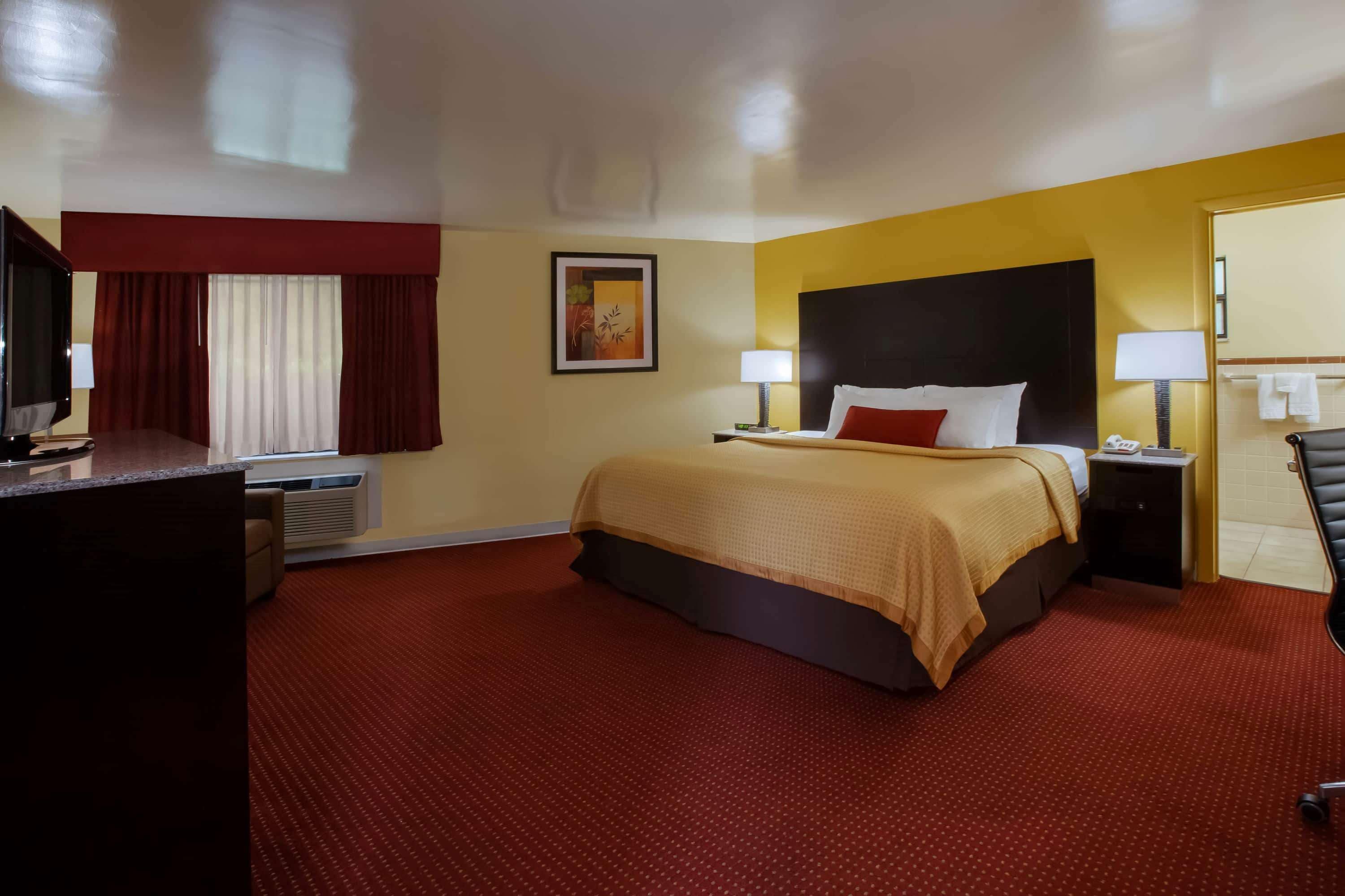 Ein Hotelzimmer mit einem großen Bett, zwei Nachttischen, einem Schreibtisch mit Computer, einem Fernseher und einem fensterbepflanzten Fenster.