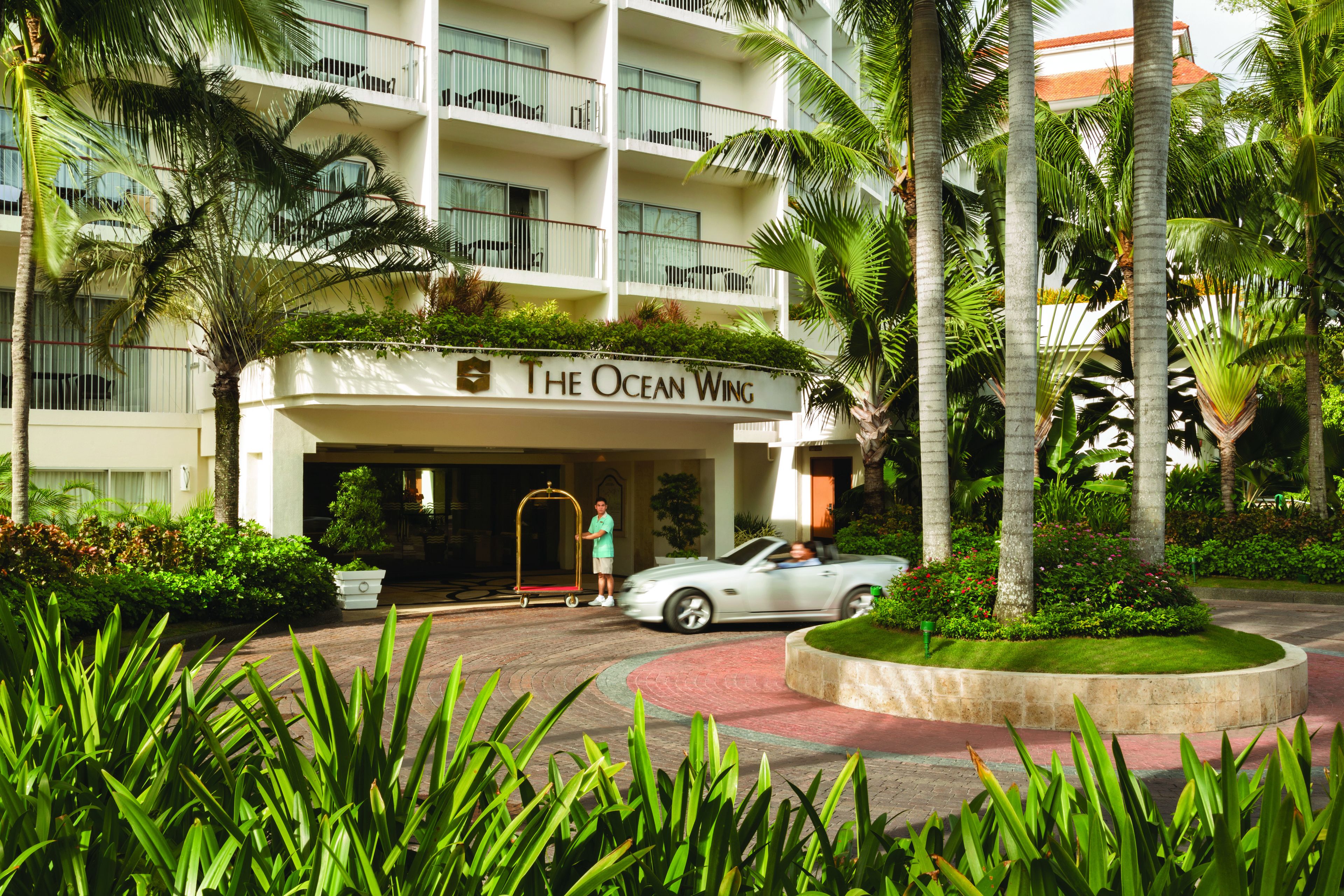 A hotel with a sign reading "The Ocean Wing," featuring a multi-story building, palm trees, and a paved driveway.