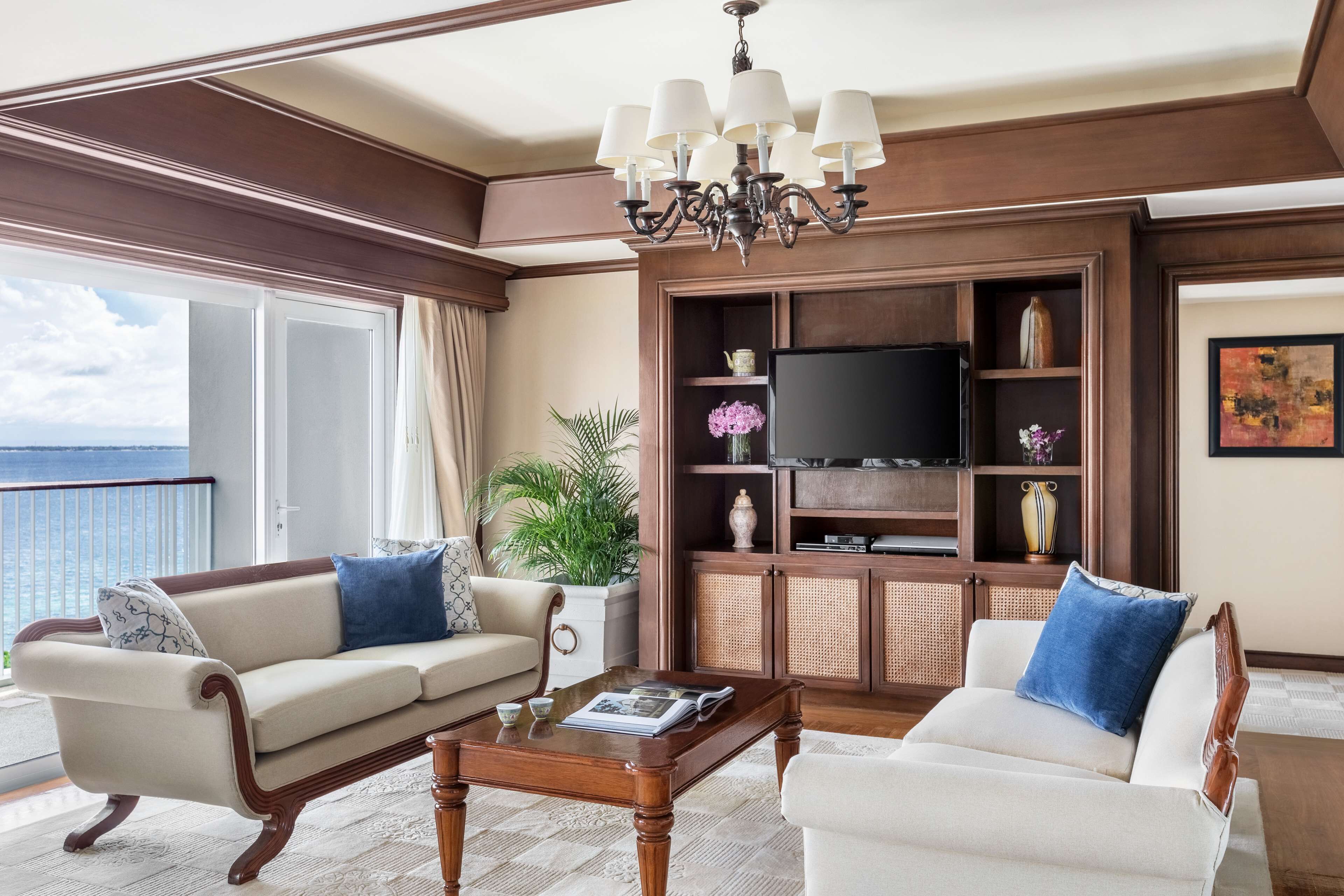 A spacious living room with a large window, a chandelier, a television, and a bookshelf.