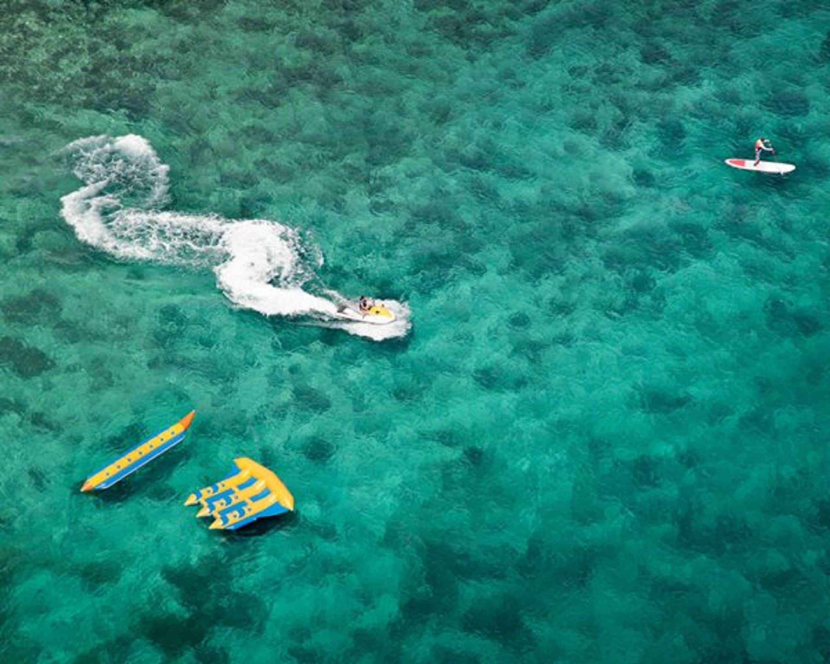Une mer turquoise aux eaux limpides, avec trois bateaux : un blanc et deux jaunes, et une personne qui rame sur le bateau blanc.