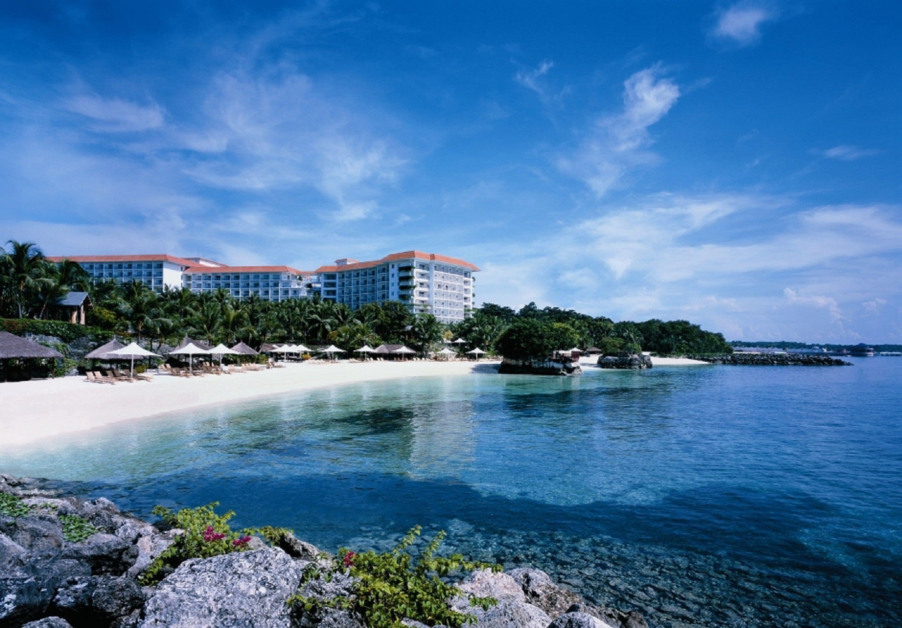 Un resort con playa, aguas azules cristalinas y un edificio grande con varios balcones.