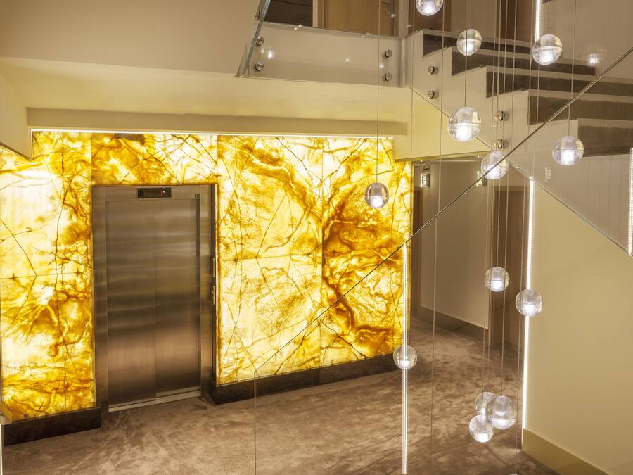 Un moderno ascensor con pared de mármol dorado, puerta metálica y escalera con luces esféricas colgantes.