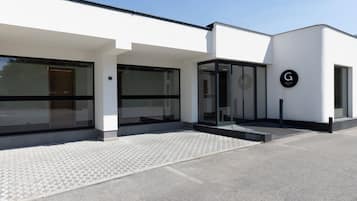 Une entrée d’hôtel moderne avec une façade en verre, un logo « G » bien visible et un chemin pavé menant à l’entrée.