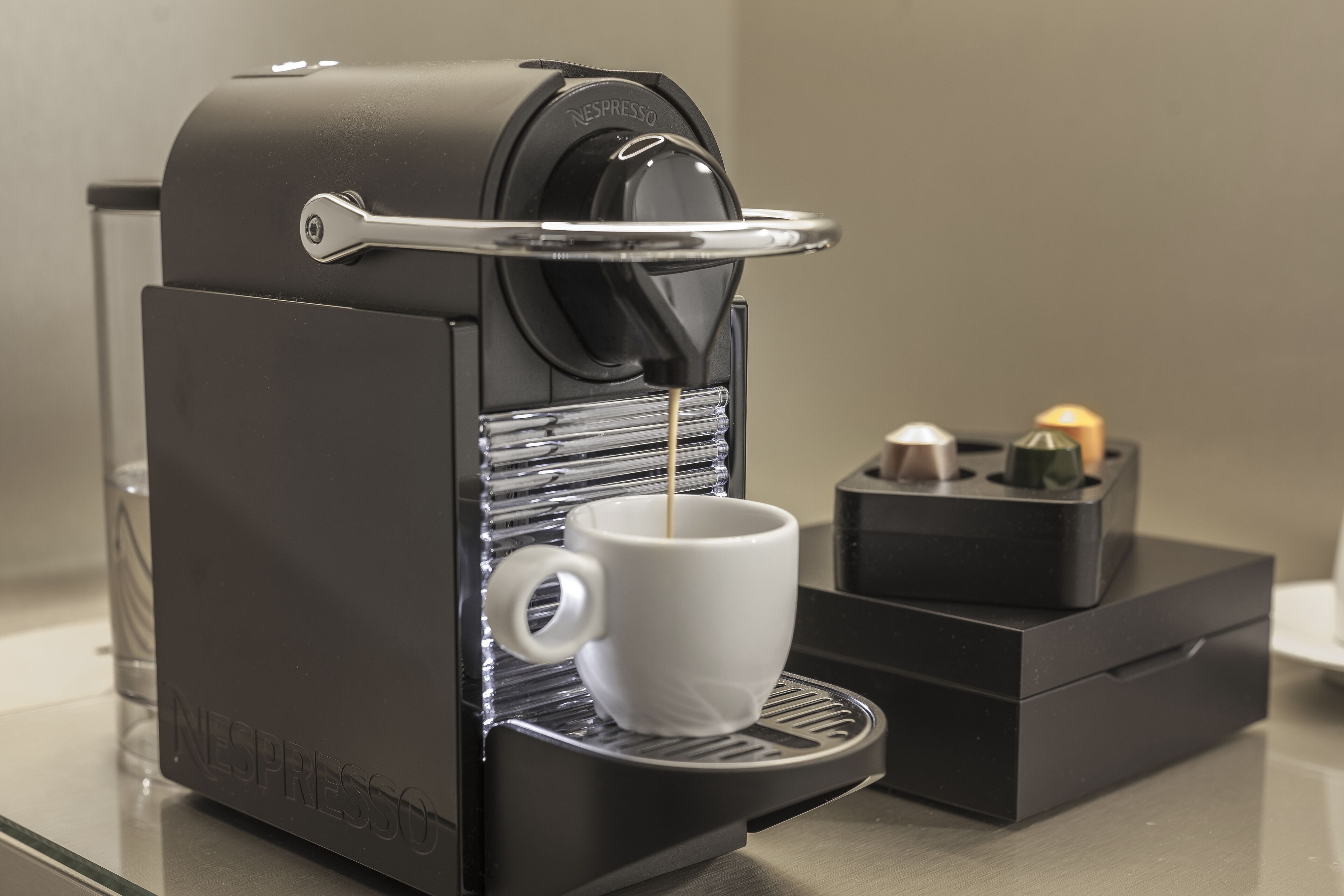 Une machine à café Nespresso avec une tasse placée sous le bec verseur, un verre d’eau et une petite boîte contenant une bougie.