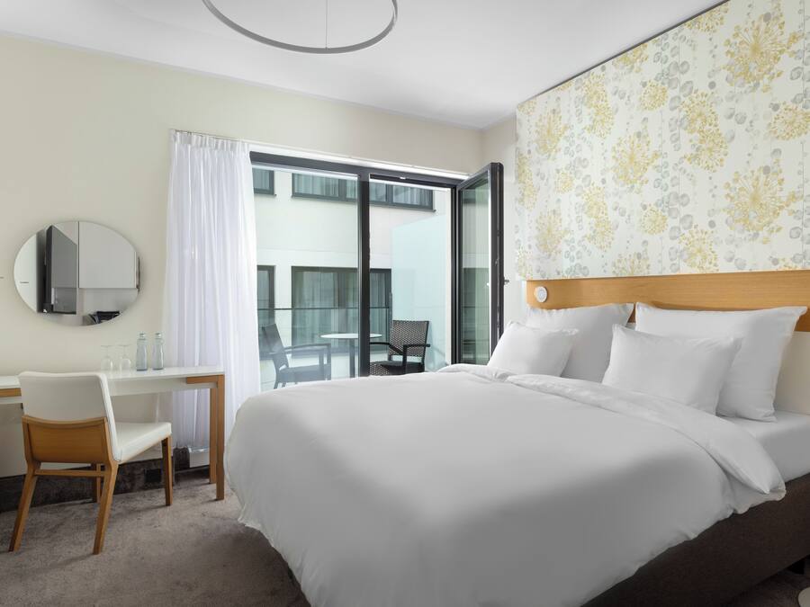 A hotel room with a large bed, a desk with a chair, a round mirror, and a window with a view of the outside.