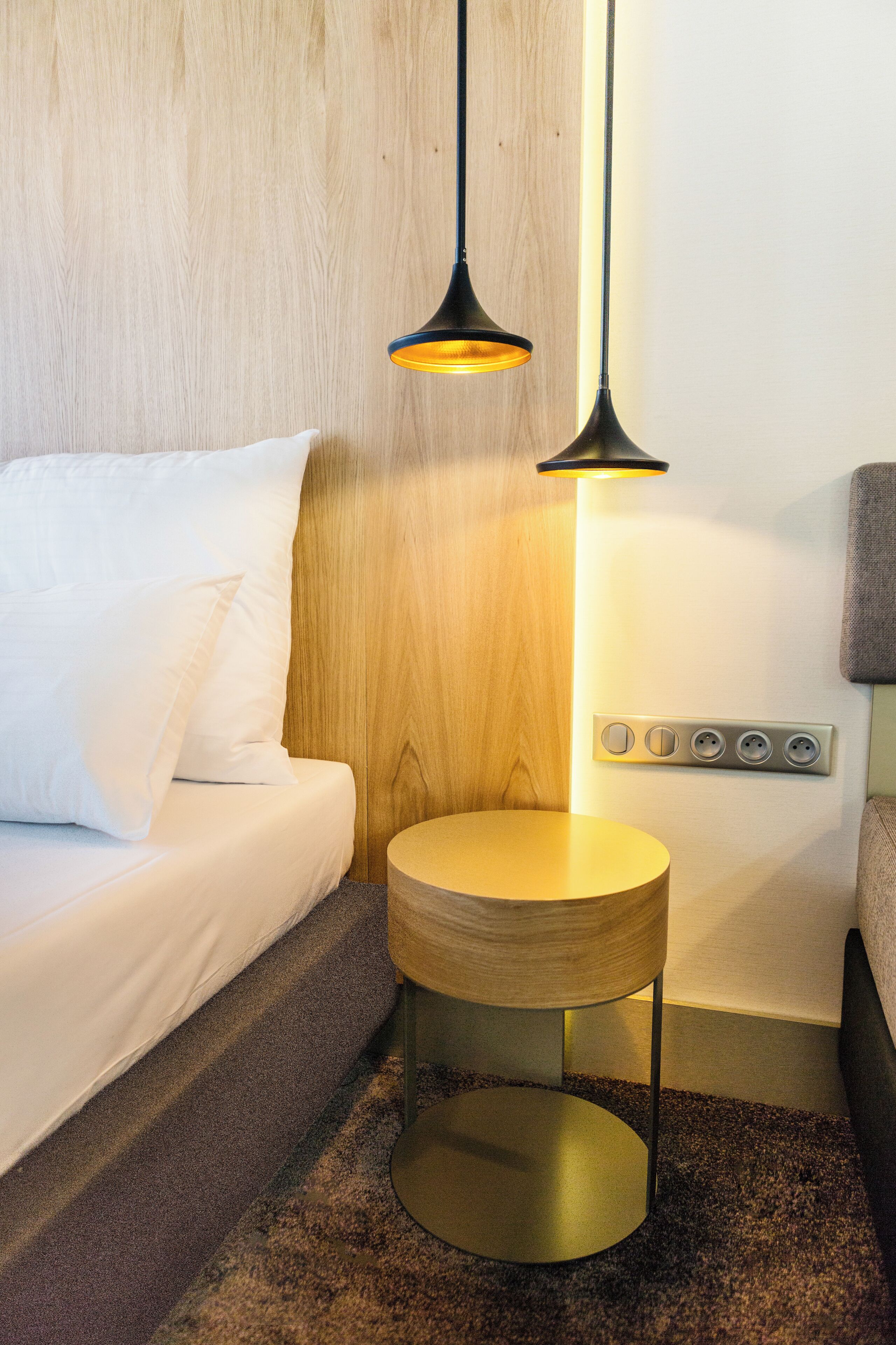 A neatly arranged hotel room with a wooden headboard, a round wooden side table, and a bed with white bedding.