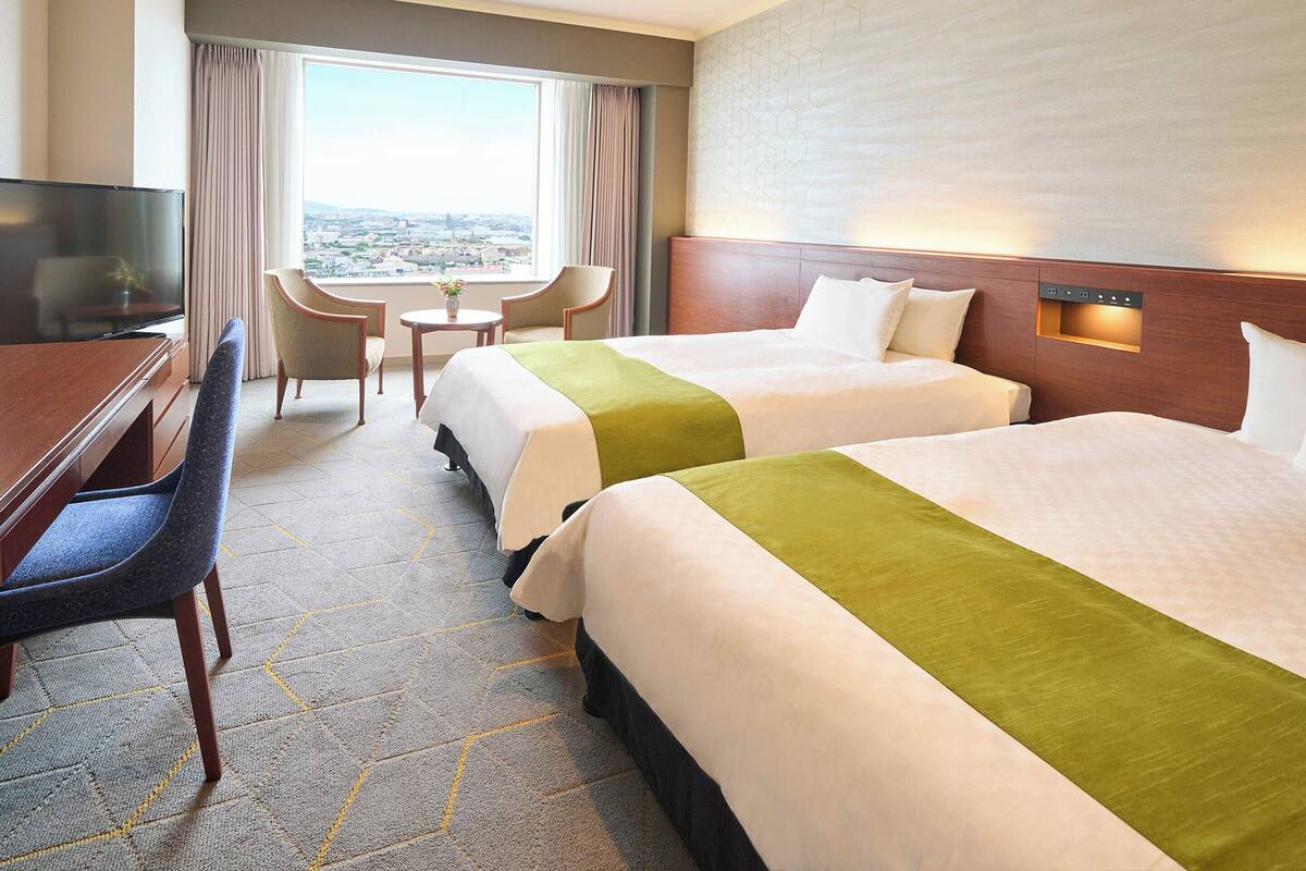 Ein Hotelzimmer mit zwei Betten, einem Schreibtisch, einem Sessel, einem Fernseher und Blick auf die Stadt.