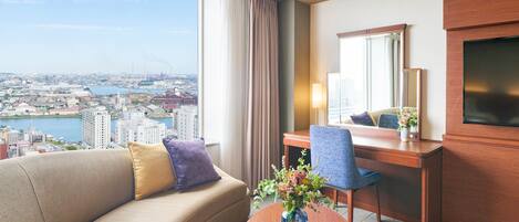 A hotel room with a sofa, a desk, a chair, a TV, and a view of a cityscape.