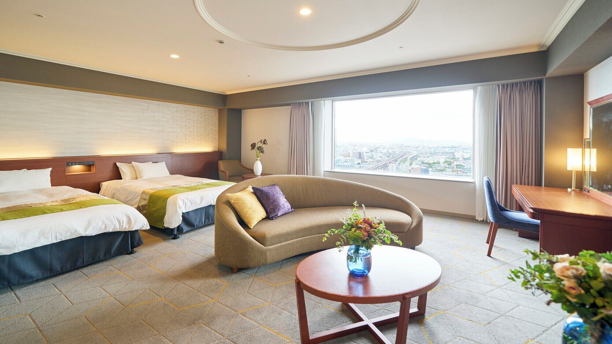 A hotel room with two beds, a sofa, a desk, and a view of the city.