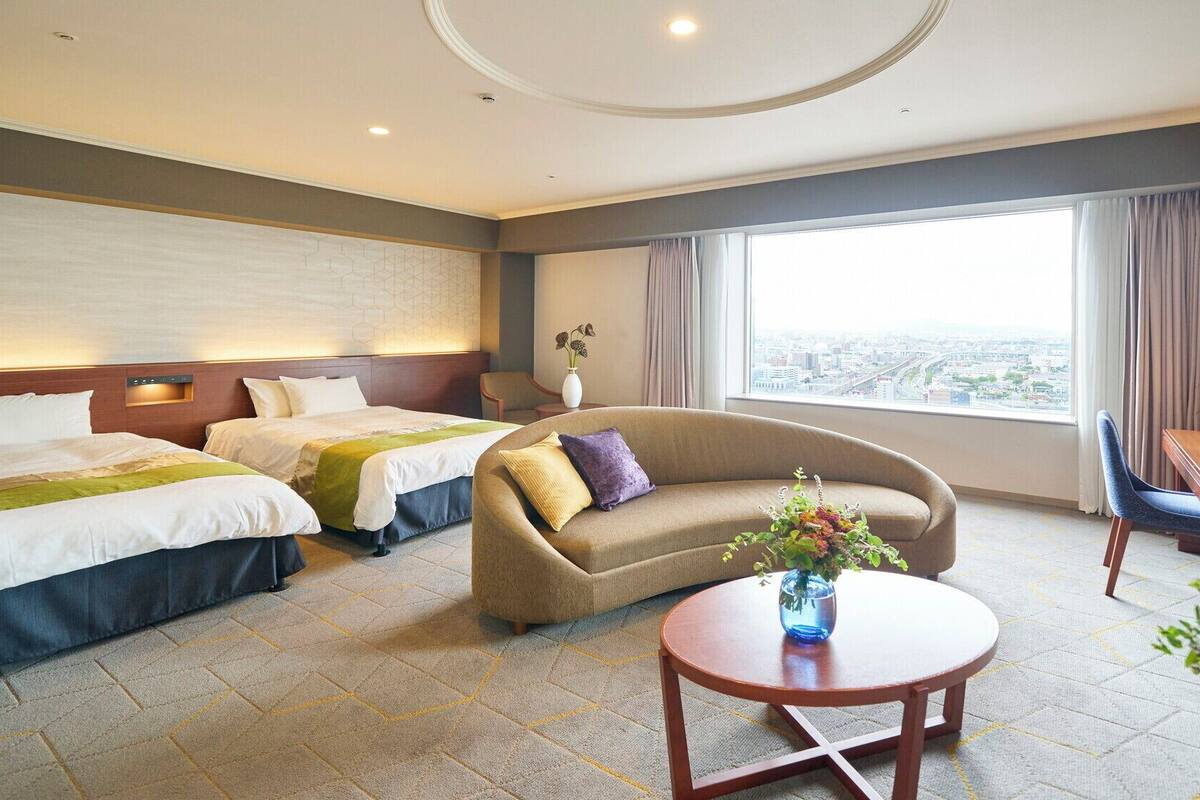 Ein Hotelzimmer mit zwei Betten, einem Sofa, einem Schreibtisch und Blick auf die Stadt.