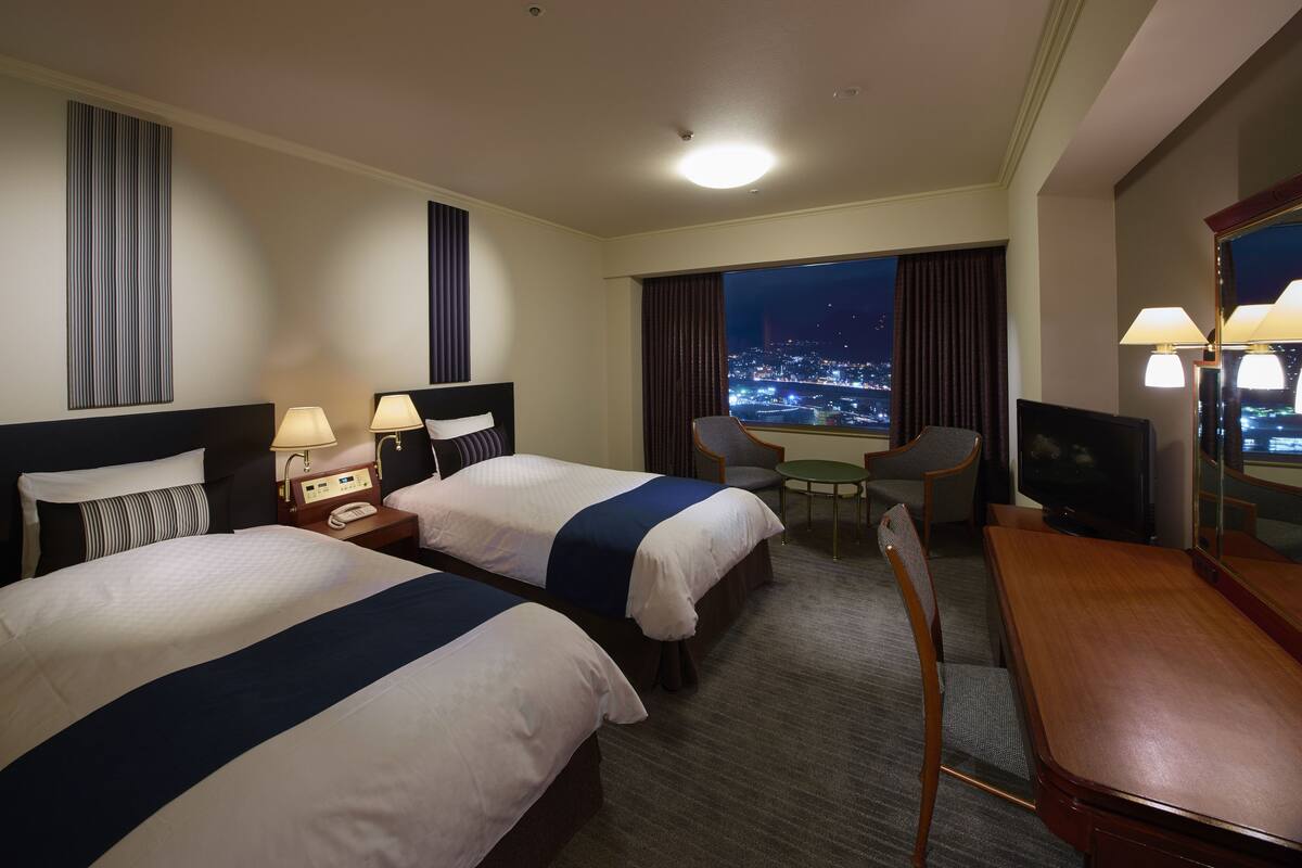 Ein Hotelzimmer mit zwei Betten, einem Schreibtisch, einem Sessel, einem Fernseher und Blick auf die beleuchtete Stadt.