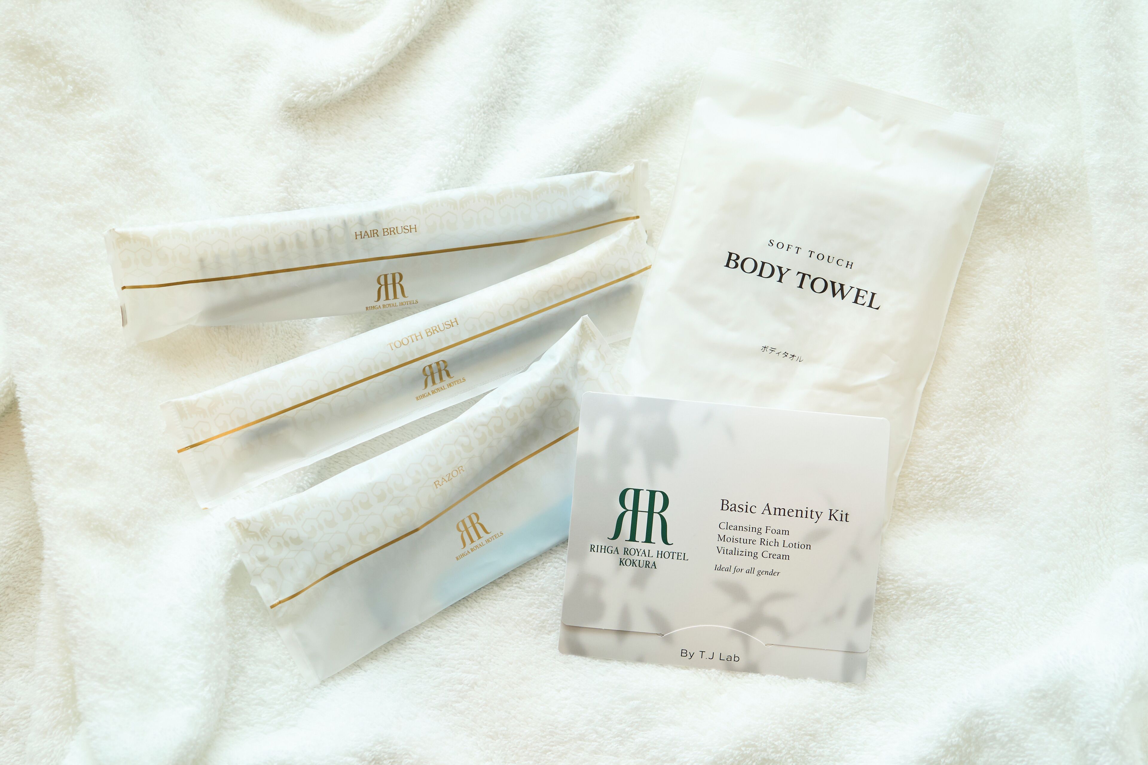 A set of white towels with gold trim and a branded logo, a soft touch body towel, and a basic amenity kit with a branded logo.