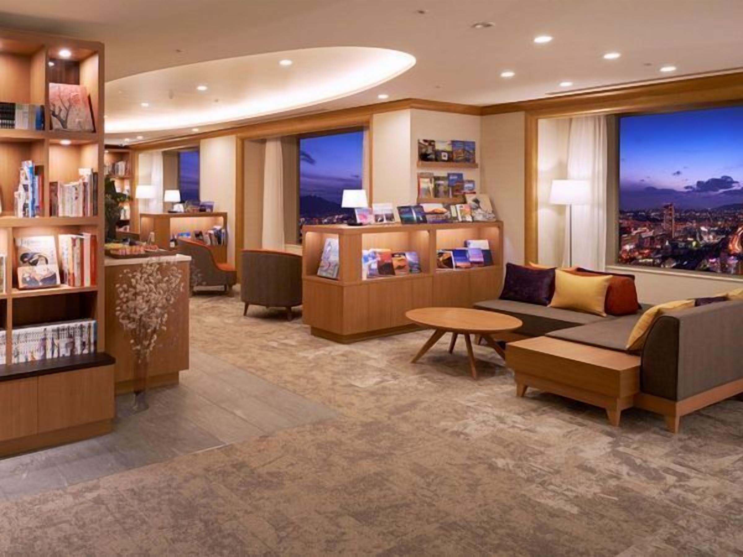 A modern hotel lobby with a seating area, bookshelves, and large windows offering a city view.