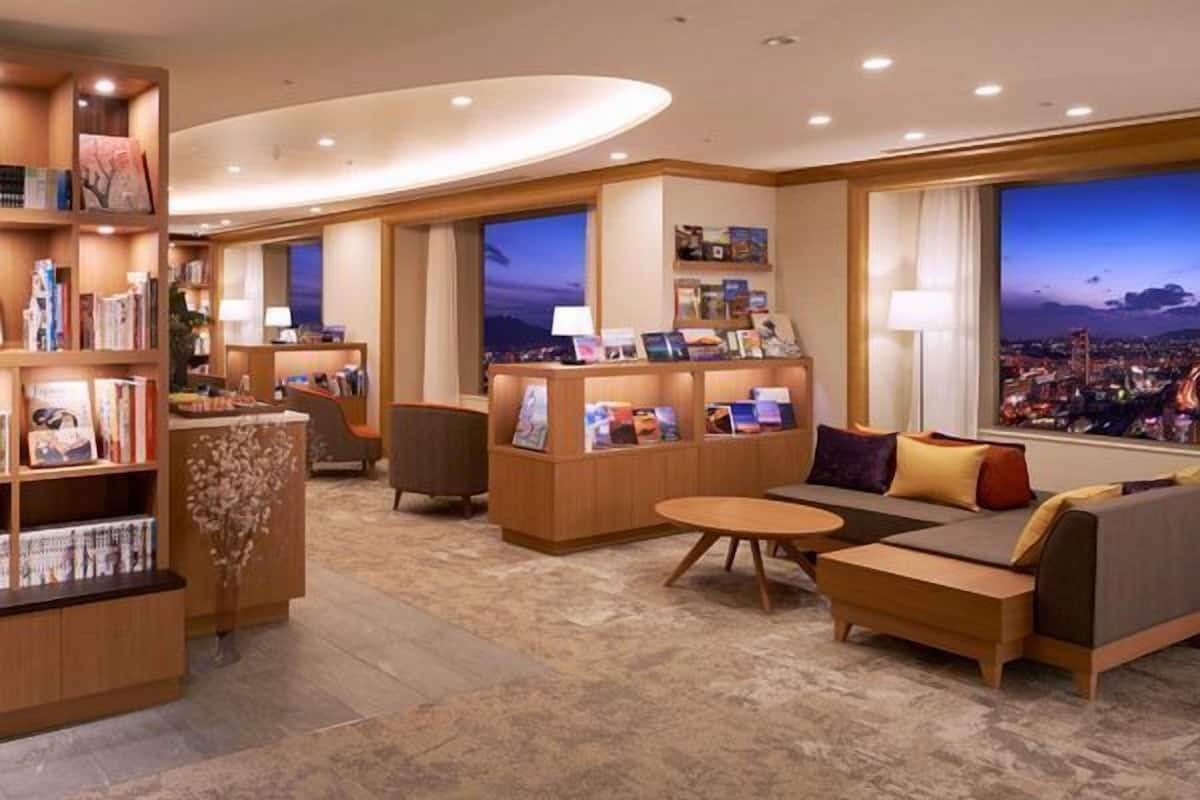 A modern hotel lobby with a seating area, bookshelves, and large windows offering a city view.