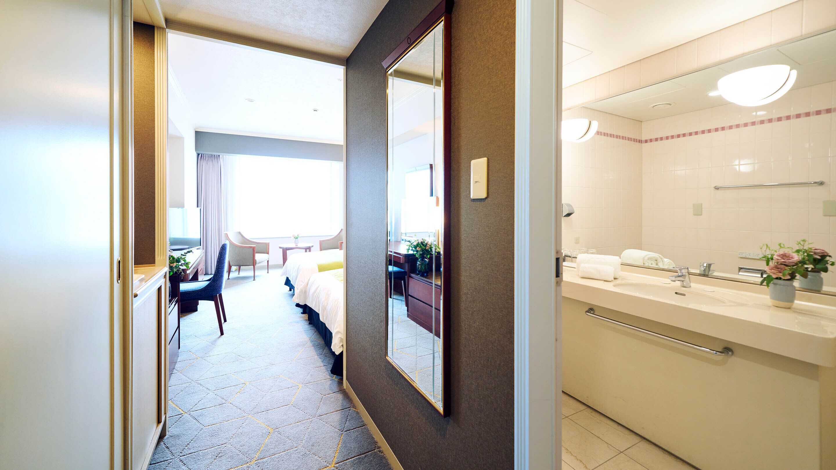Habitación de hotel con cama, escritorio, silla, mesita y baño con lavamanos y espejo.