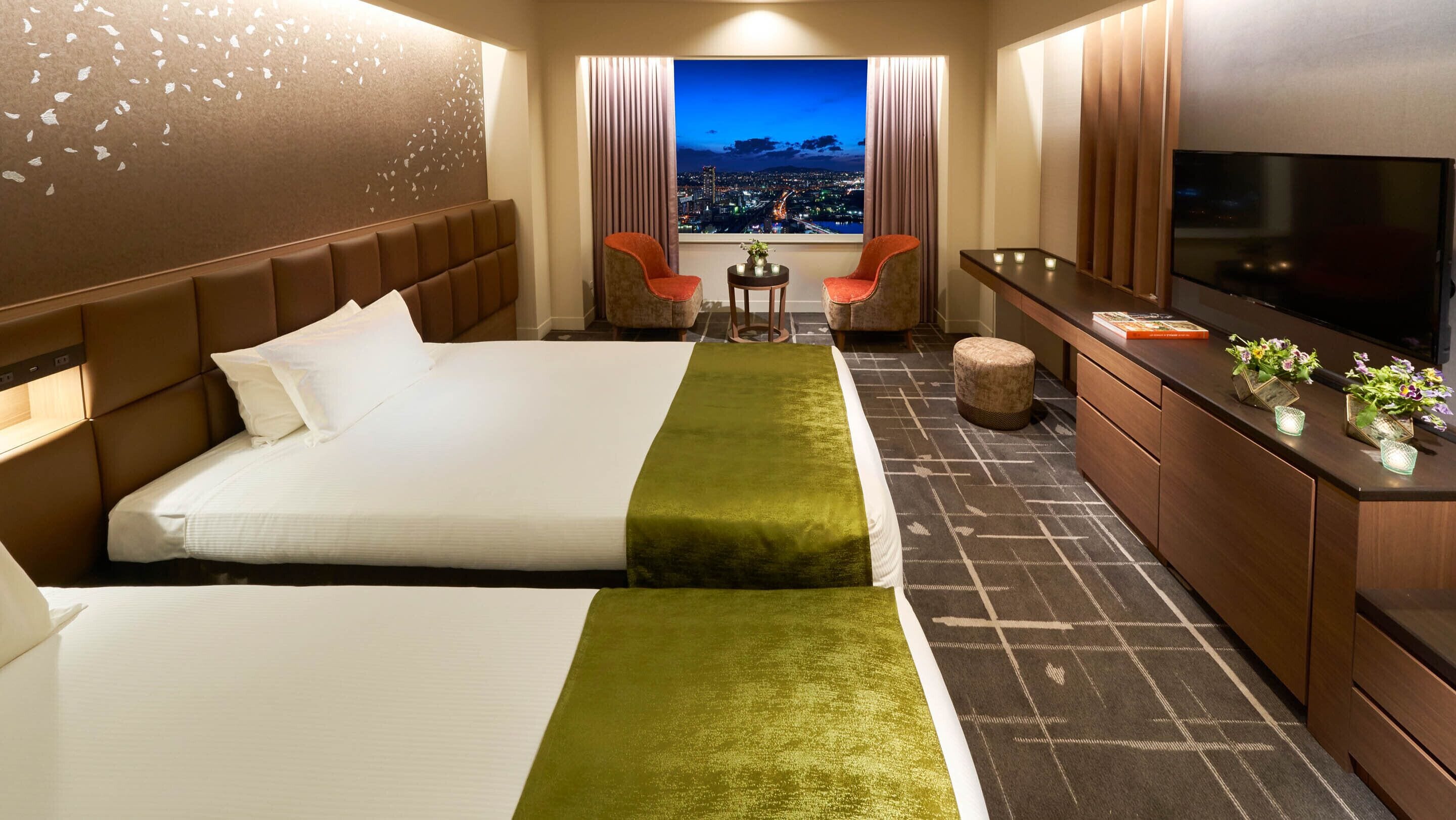 Een hotelkamer met twee bedden, een televisie, een klein tafeltje met bloemen en uitzicht op een nachtelijke stadsgezicht.