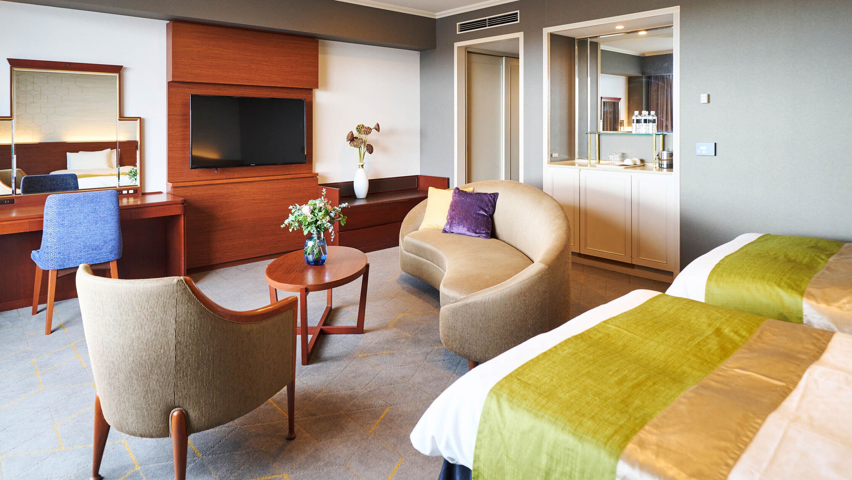 Habitación de hotel con cama, dos sillones, sofá, escritorio con silla, televisor, florero con flores y espejo.