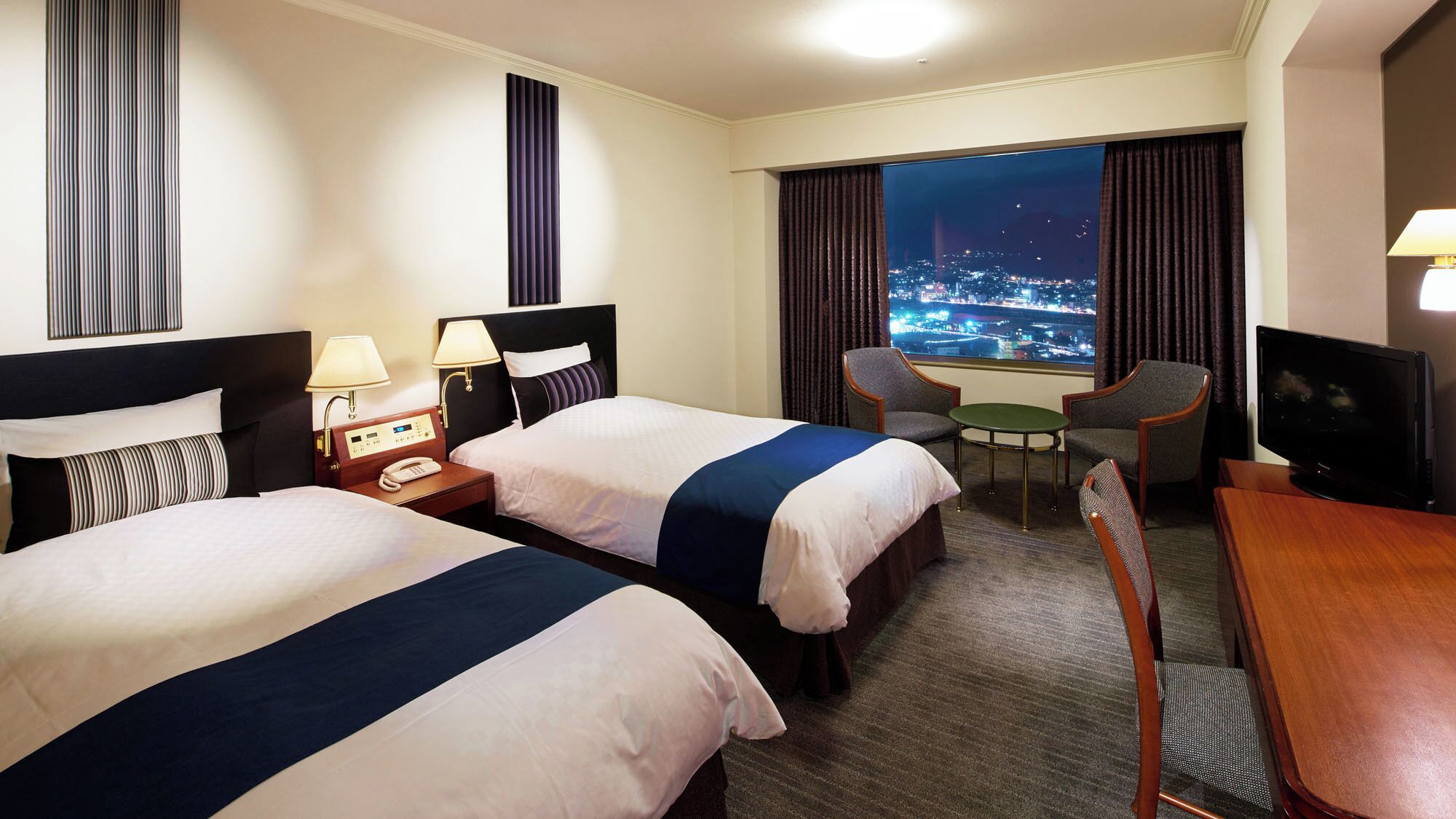 A hotel room with two beds, a desk with a TV, a chair, a small table, and a view of a cityscape at night.