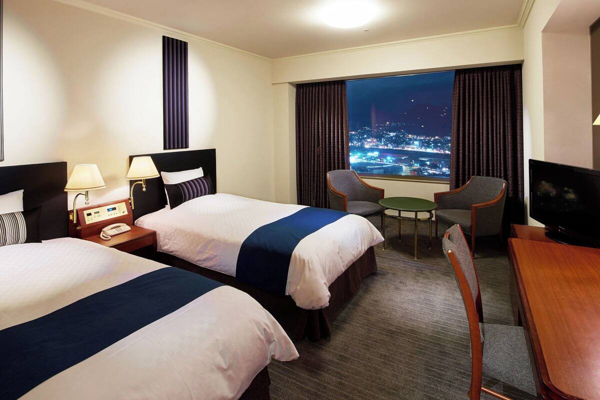 Ein Hotelzimmer mit zwei Betten, einem Schreibtisch mit Fernseher, einem Sessel, einem kleinen Tisch und Blick auf eine nächtliche Skyline.