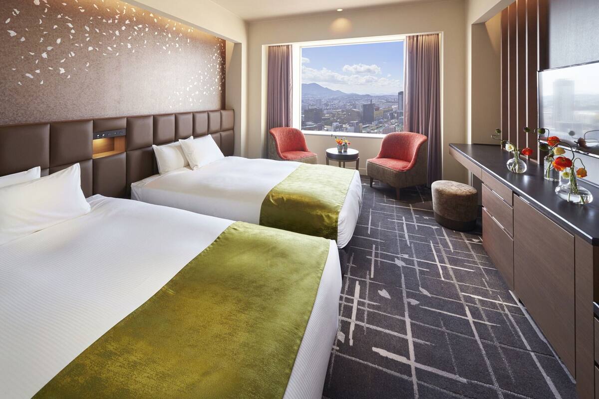 Ein Hotelzimmer mit zwei Betten, Blick auf eine Stadtansicht, einem kleinen Tisch und einer Sitzecke mit Stühlen.