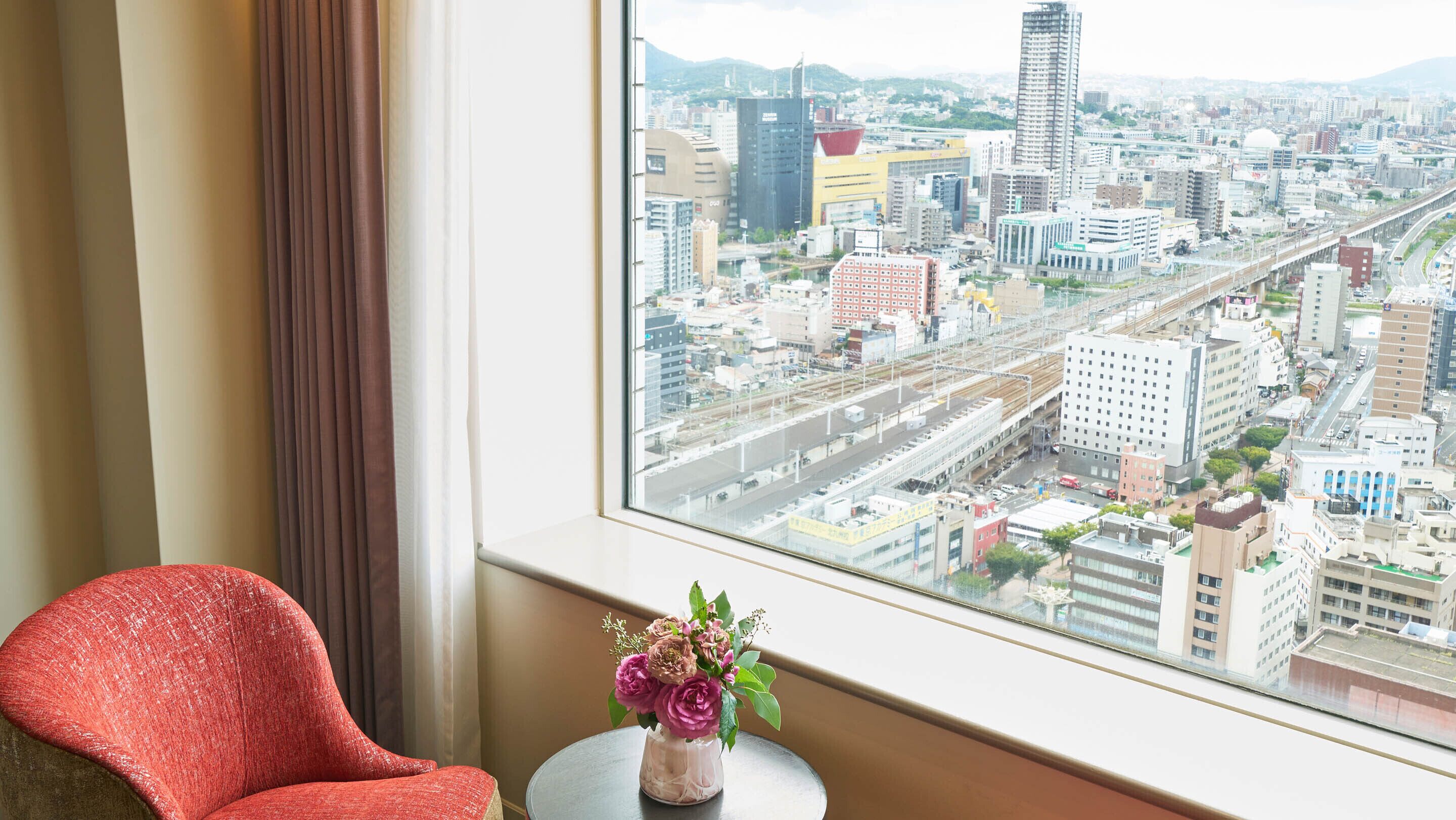 Une pièce avec vue sur le paysage urbain, visible à travers une fenêtre, un fauteuil rouge et un vase rempli de fleurs posé sur une petite table.