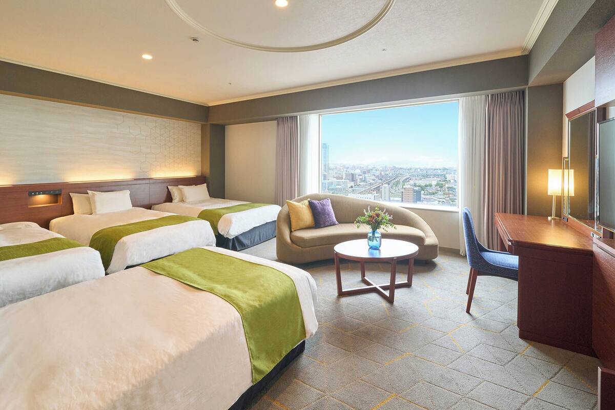 Ein Hotelzimmer mit drei Betten, einem Schlafsofa, einem Schreibtisch und einem großen Fenster mit Blick auf die Stadt.