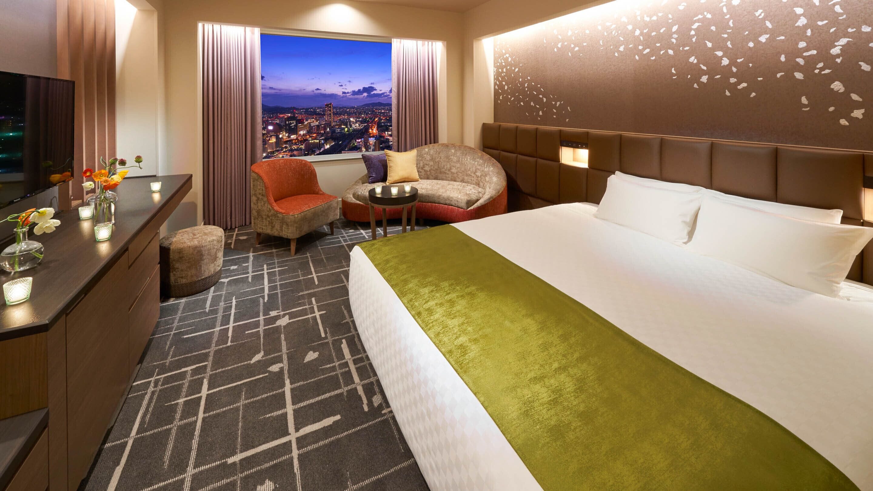 A hotel room with a large bed, a desk with a TV, a chair, a small table with candles, and a view of a cityscape at night.