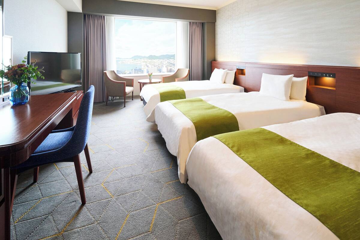 Ein Hotelzimmer mit zwei Betten, einem Schreibtisch, einem Sessel und einem Fernseher, mit Blick auf die Stadt.