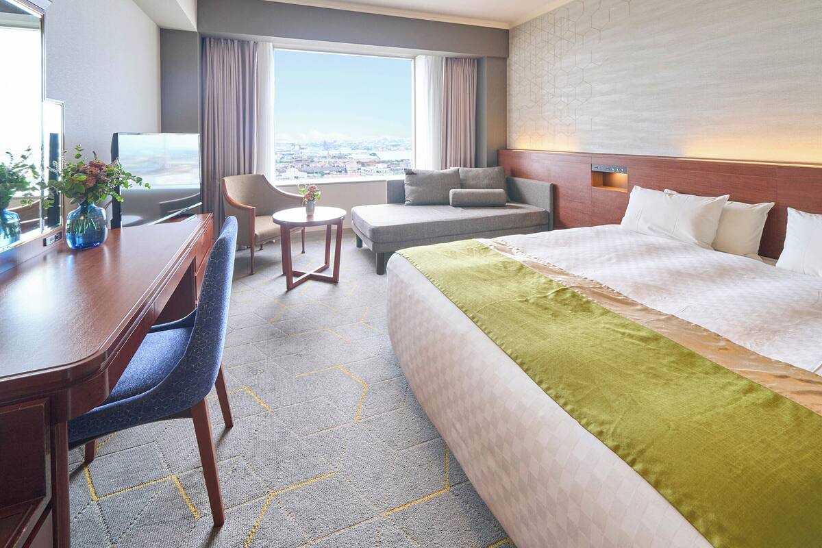 Ein Hotelzimmer mit einem großen Bett, einem Schreibtisch, einem Sessel, einem kleinen Tisch und Blick auf die Stadt durch das Fenster.