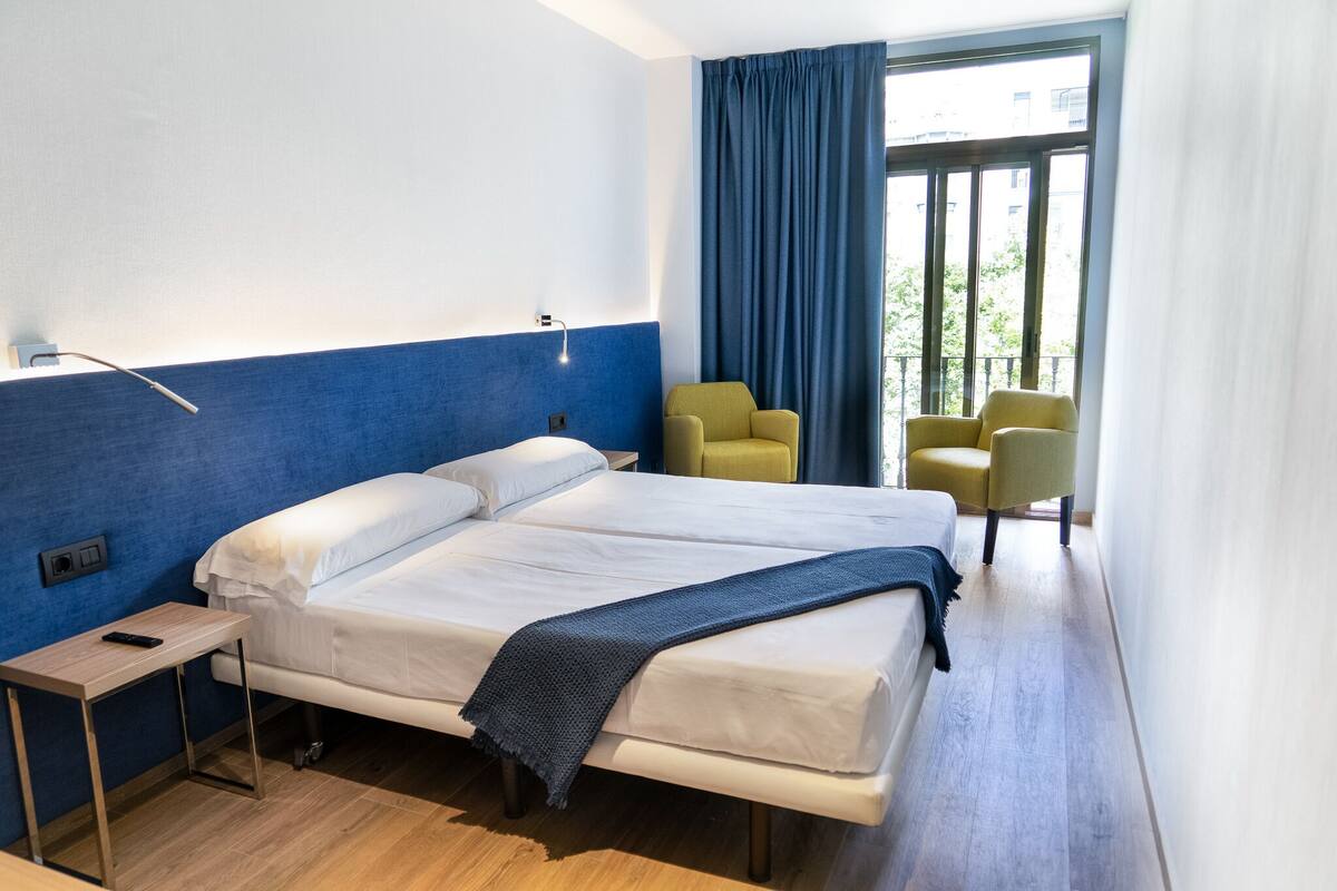 A hotel room with a bed, a bedside table with a remote control, a chair, and a view of the outdoors through a window with blue curtains.