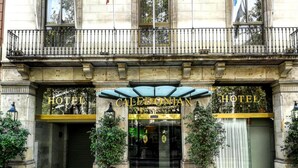 The exterior of the Hotel Caledonian with a classic stone facade, large glass entrance doors, and a balcony with a metal railing.