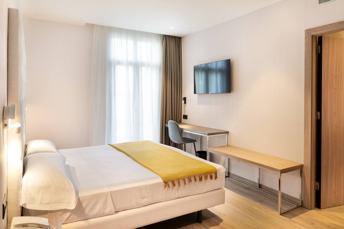 A hotel room with a bed, a desk, a chair, a TV, and a wooden door.