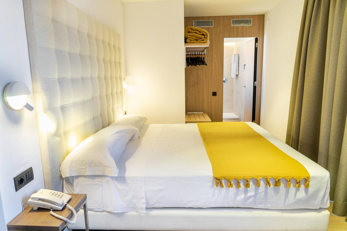 A hotel room with a bed, a bedside phone, a headboard, a yellow throw blanket, and a coat rack with a folded jacket.