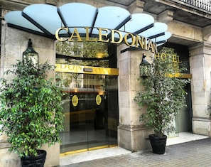 The entrance to the Caledonian Hotel with a glass facade, a prominent gold sign, and two potted plants. - Hotel Caledonian (Barcelona)