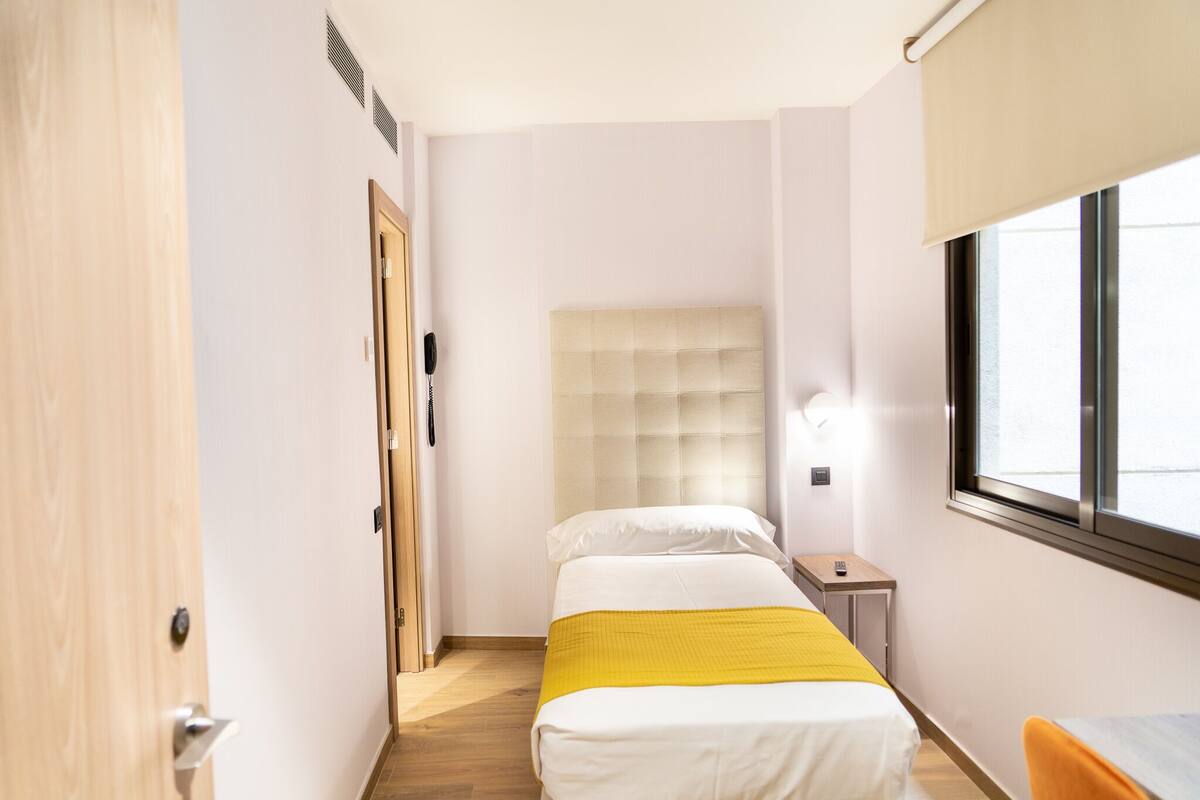 A narrow hotel room with a bed, bedside table, and a window.