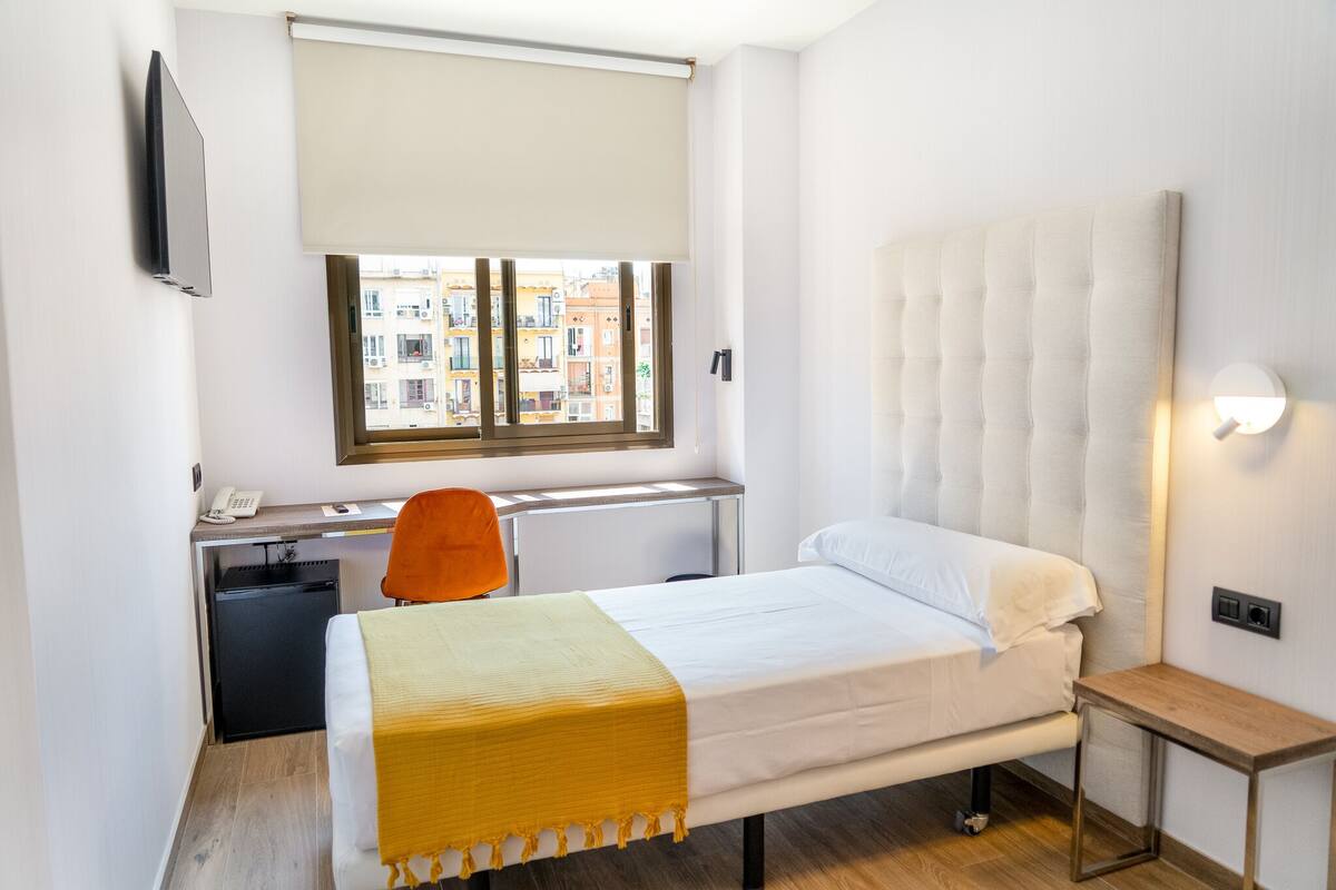 A hotel room with a bed, a desk with a chair, a small refrigerator, a window with blinds, and a wall-mounted TV.