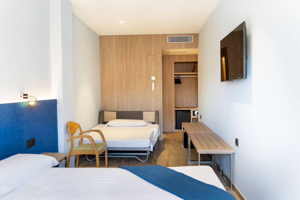 A modern hotel room with a single bed, a desk, a chair, and a TV mounted on the wall.