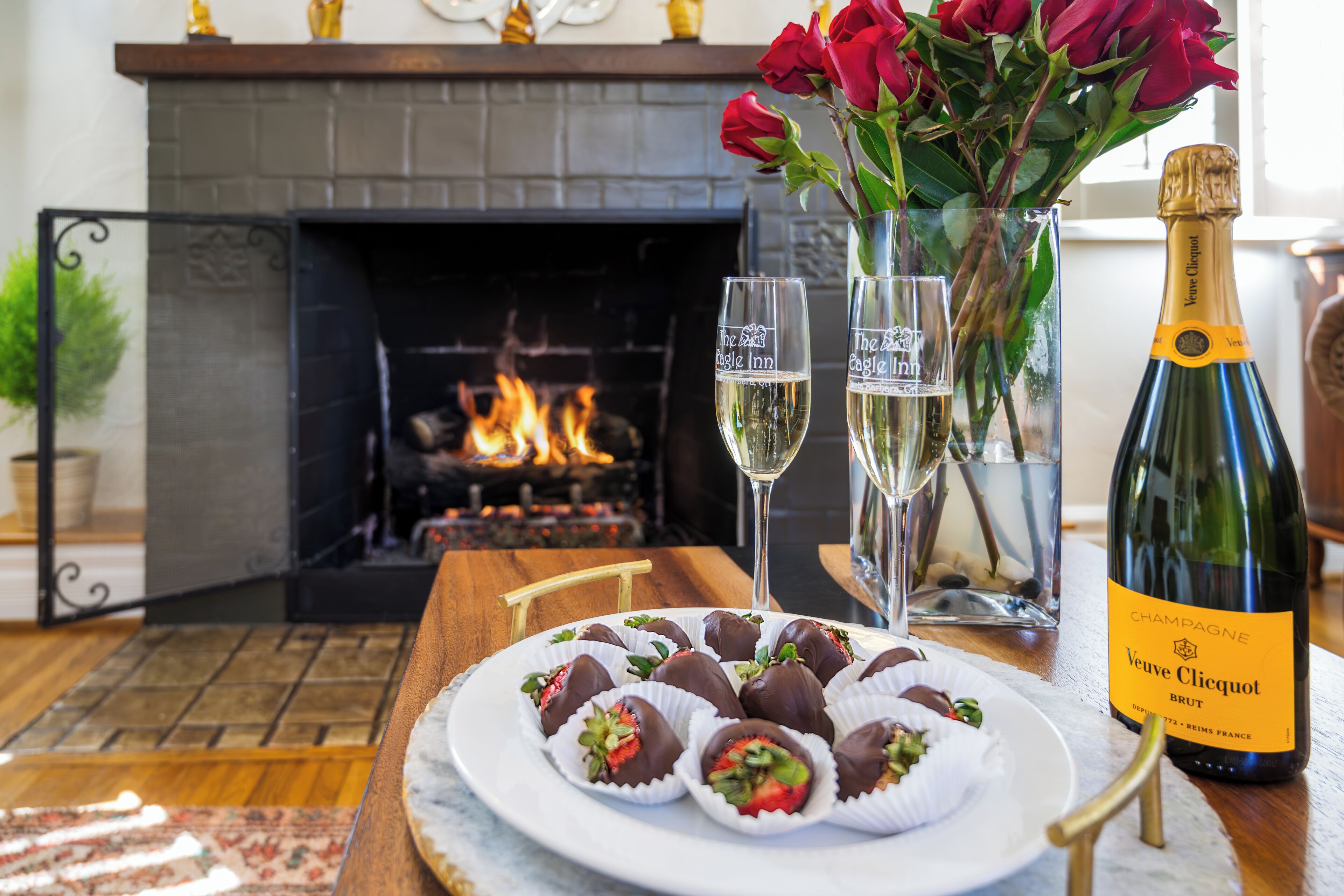 Une assiette de fraises enrobées de chocolat, deux coupes à champagne et une bouteille de Veuve Clicquot sur une table devant une cheminée.