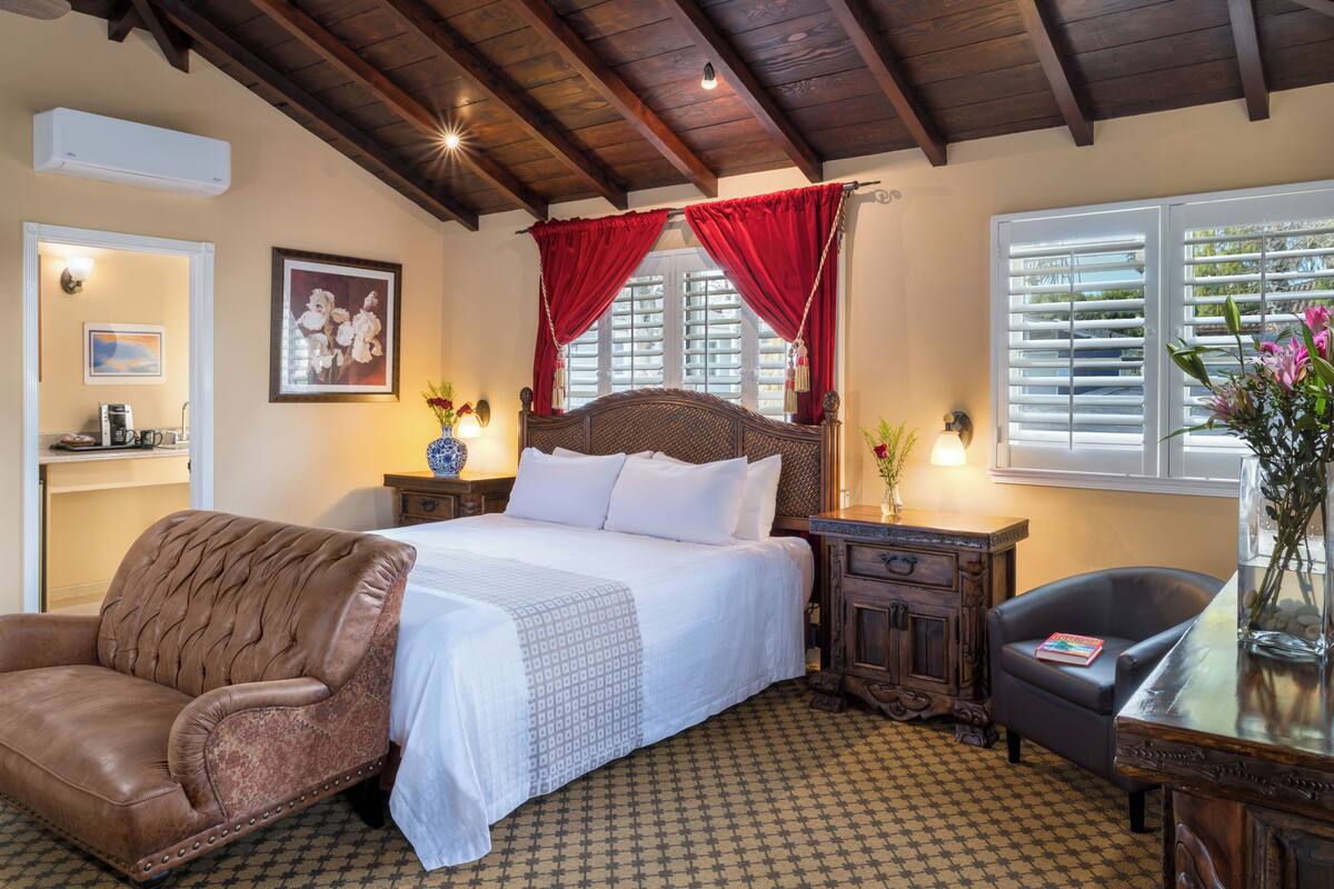 A bedroom with a wooden bed, red curtains, a leather armchair, a bedside table with a lamp, and a window with blinds.